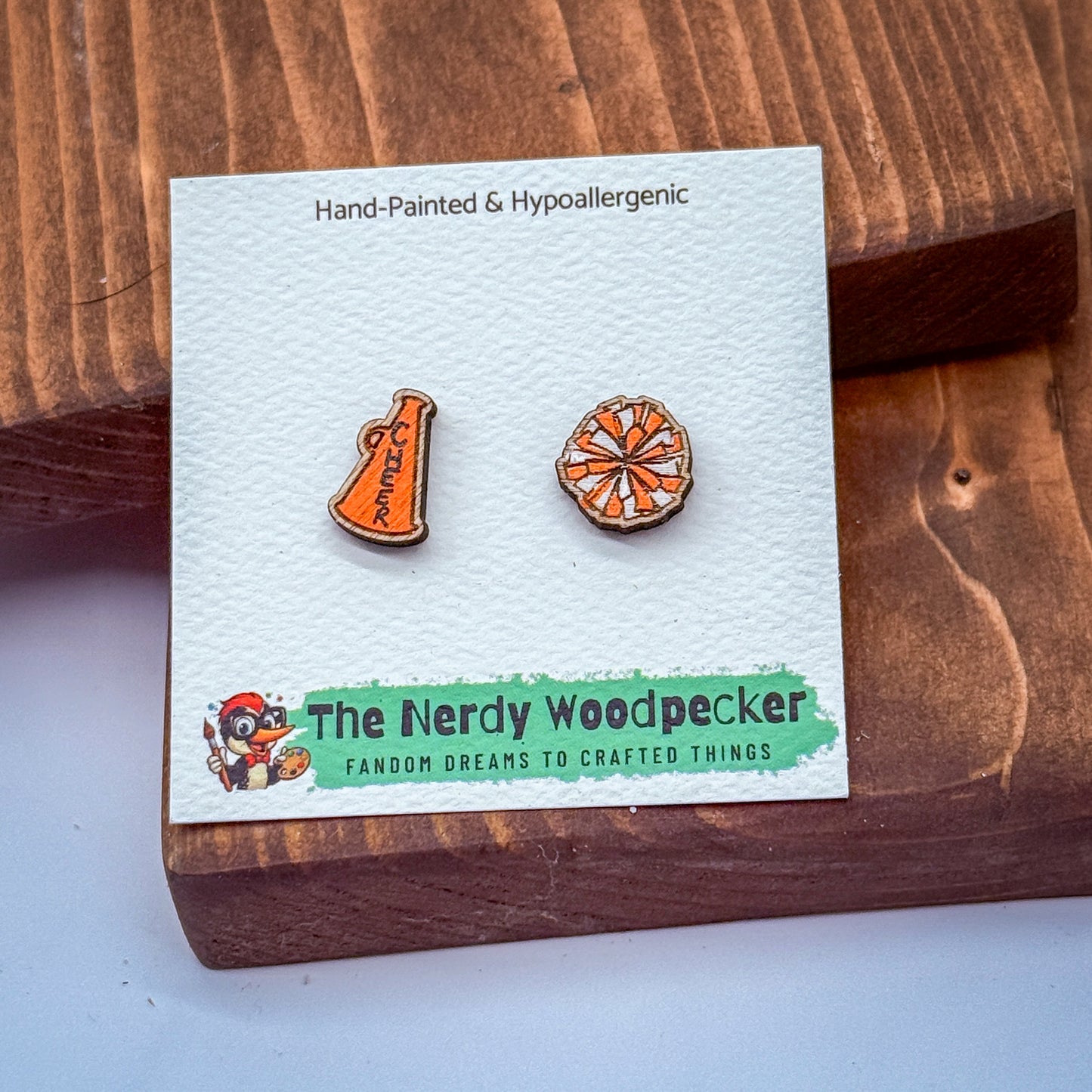 a pair of earrings, one shaped like a cheerleader and the other like a wheel, displayed on a wooden surface.