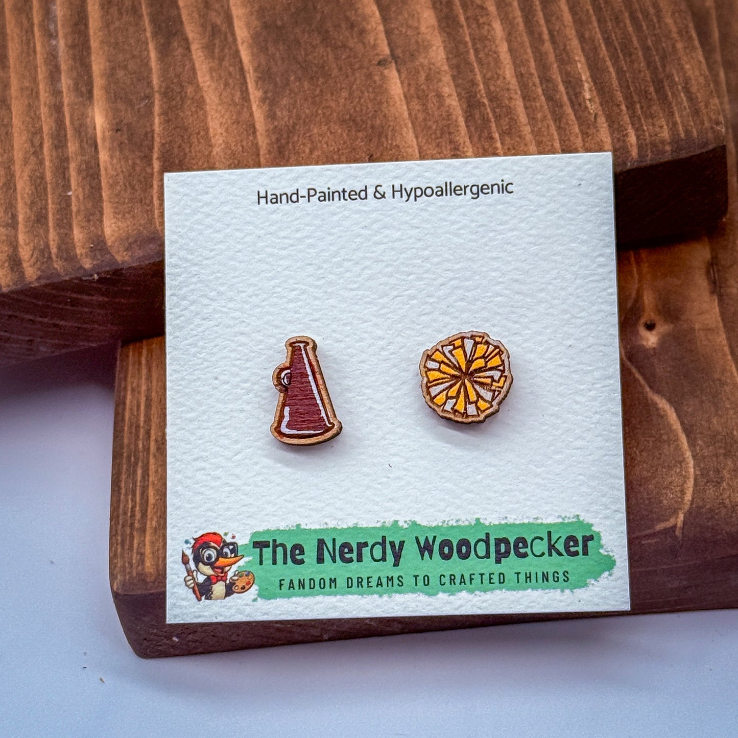 a pair of hand-painted hypoallergenic earrings, one shaped like a megaphone and the other a flower, displayed on a wooden surface.