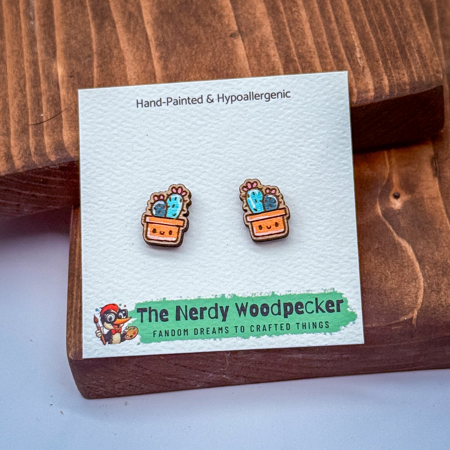 a pair of hand-painted and hypoallergenic earrings with cactus designs, displayed on a wooden surface.