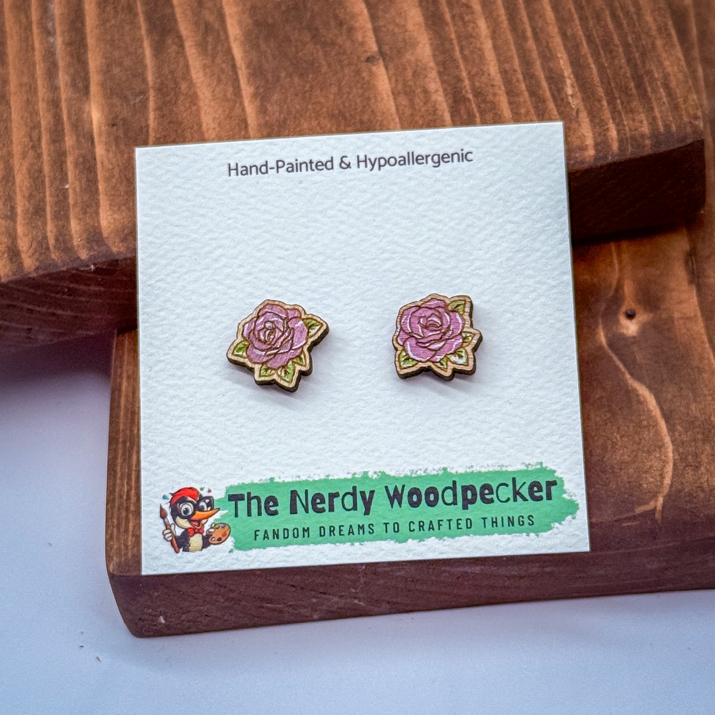 a pair of hand-painted and hypoallergenic earrings displayed on a wooden surface. The earrings are part of a product from The Nerdy Woodpecker, a fan of crafting and creating things.