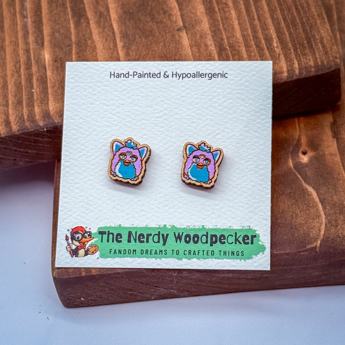 a pair of earrings in the shape of a cartoon character, placed on a wooden surface. The earrings are described as hand-painted and hypoallergenic, and they are part of a product from "The Nerdy Woodpecker" brand.