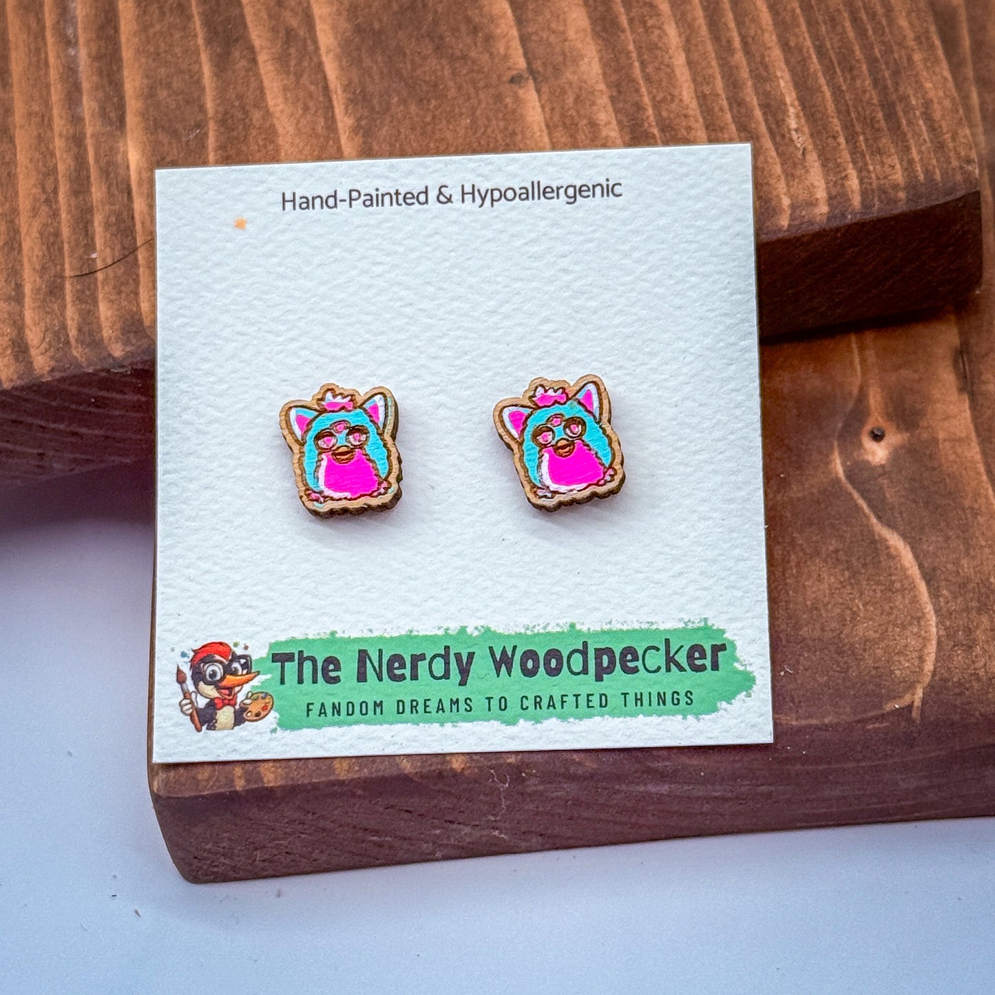 a pair of earrings in the shape of a pink and blue bird, placed on a white card with text and a cartoon character.
