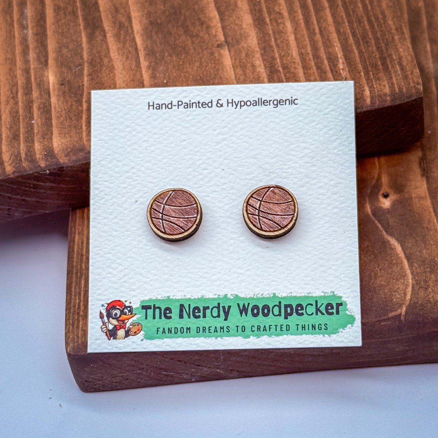 a pair of hand-painted and hypoallergenic earrings from The Nerdy Woodpecker, a fan of crafting and creating things. The earrings are displayed on a wooden surface, and the packaging features a cartoon character and text that describes the earrings as "Famous for their unique design and craftsmanship".