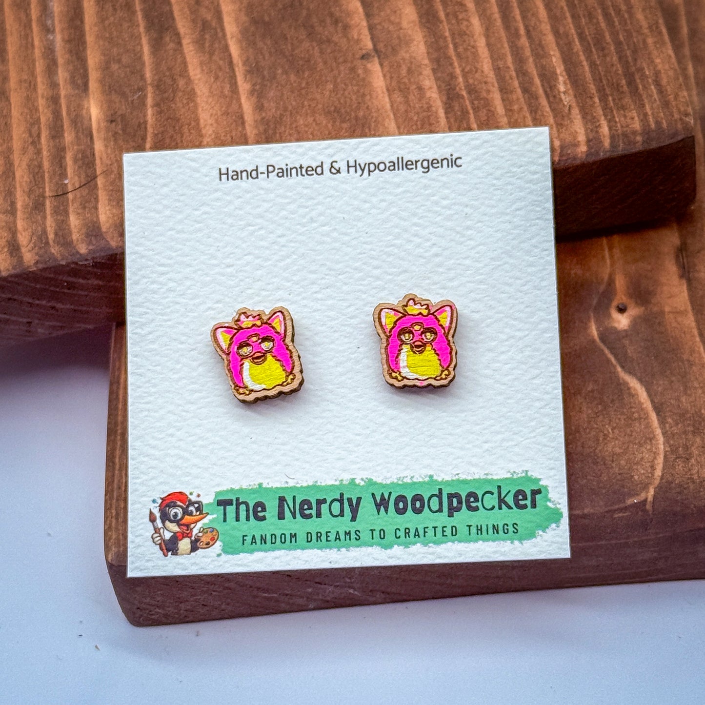 a pair of hand-painted earrings with a pink and yellow design, placed on a white card with text and a cartoon character.