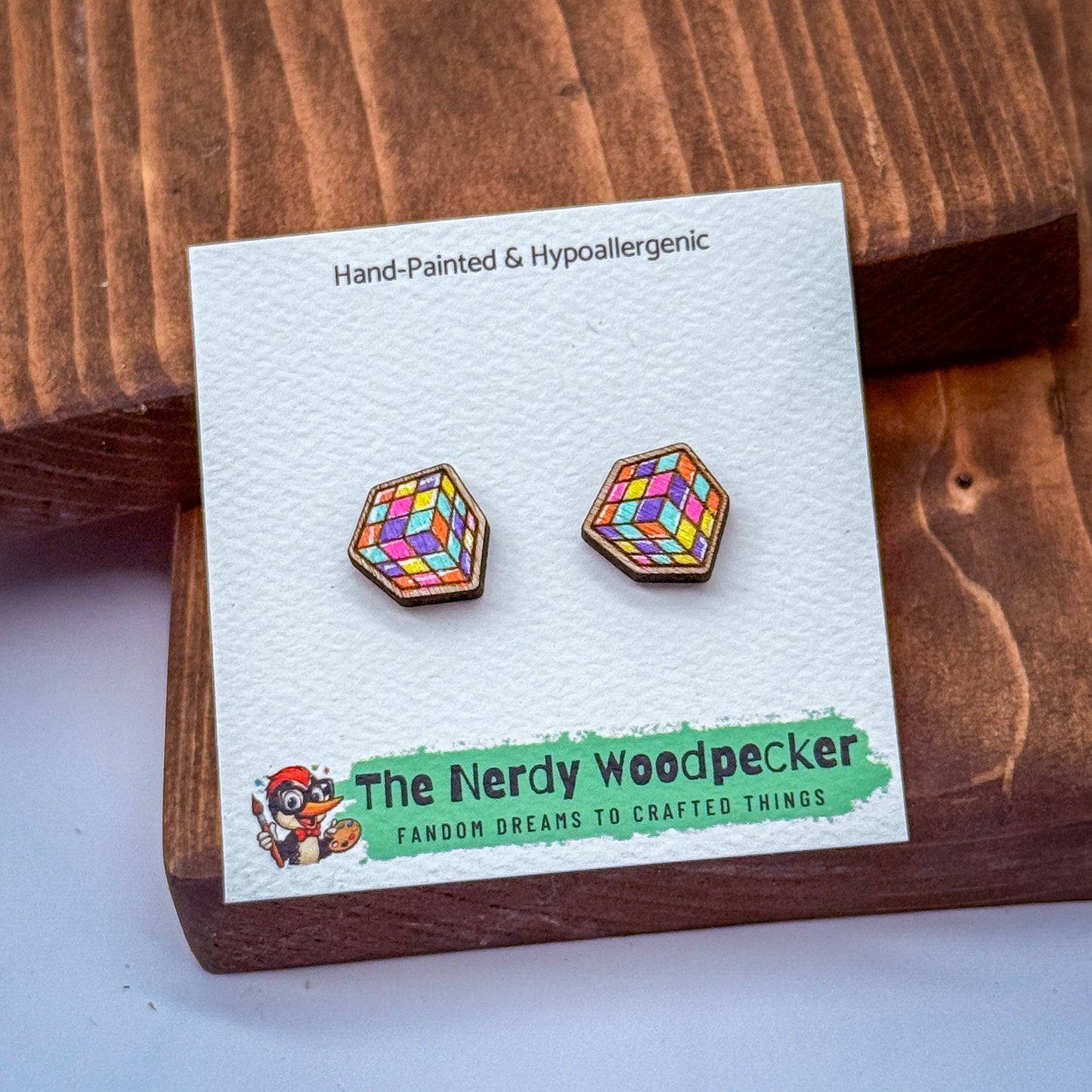 a pair of earrings with a colorful geometric design, placed on a wooden surface. The earrings are presented in a white card with text and a logo.