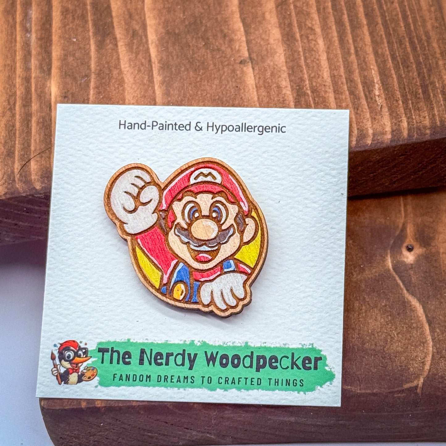 A hand-painted and hypoallergenic pin of Mario, the iconic character from the Super Mario video game franchise, is displayed on a wooden surface.