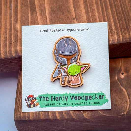 A hand-painted wooden pin depicting a character from the Star Wars franchise, specifically the character Baby Yoda, holding a green alien-like creature. The pin is displayed on a white card with text and a graphic.