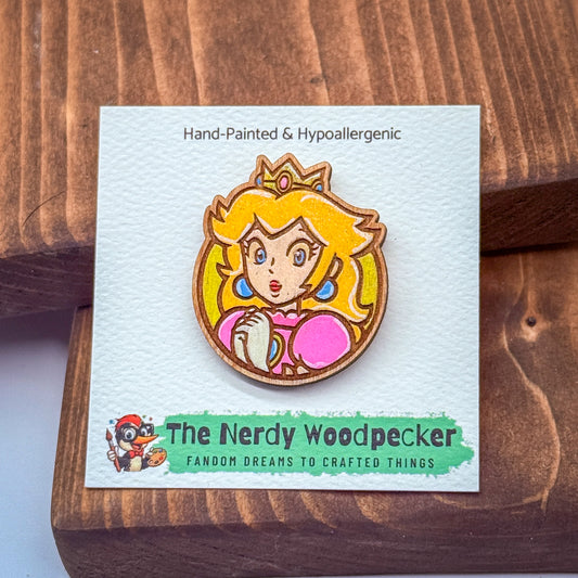 A hand-painted wooden pin featuring a cartoon character, likely a princess or fairy, with a pink dress and crown. The pin is displayed on a white card with text and a logo.
