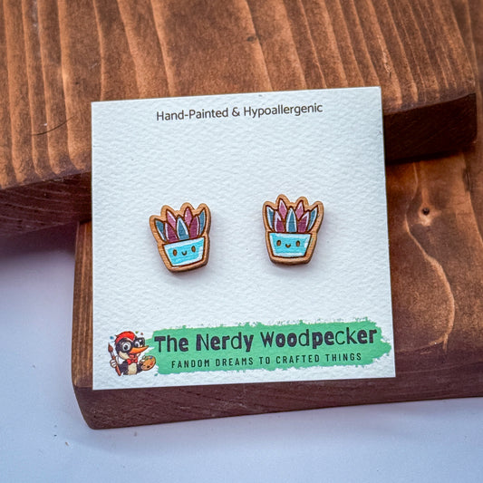 a pair of earrings with a hand-painted design, placed on a wooden surface. The earrings are labeled as hypoallergenic and are part of a collection by The Nerdy Woodpecker, which features whimsical, hand-painted designs.
