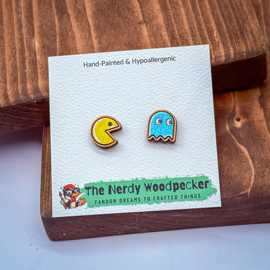 two earrings, one shaped like Pac-Man and the other like a ghost, displayed on a wooden surface.
