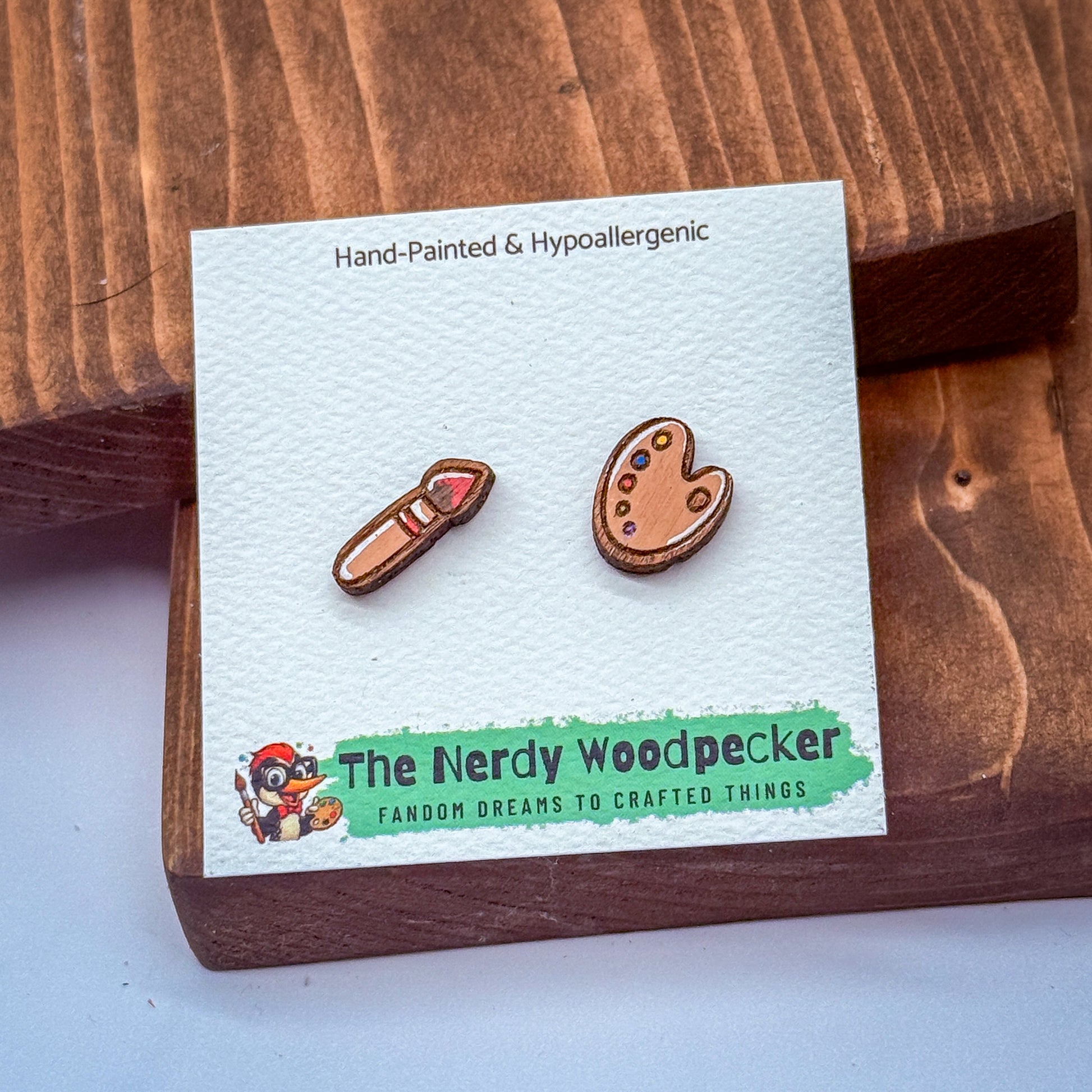 a pair of hand-painted hypoallergenic earrings, with a wooden background and a green and white label that reads "The Nerdy Woodpecker FANDOM DREAMS TO CRAFTED THINGS".