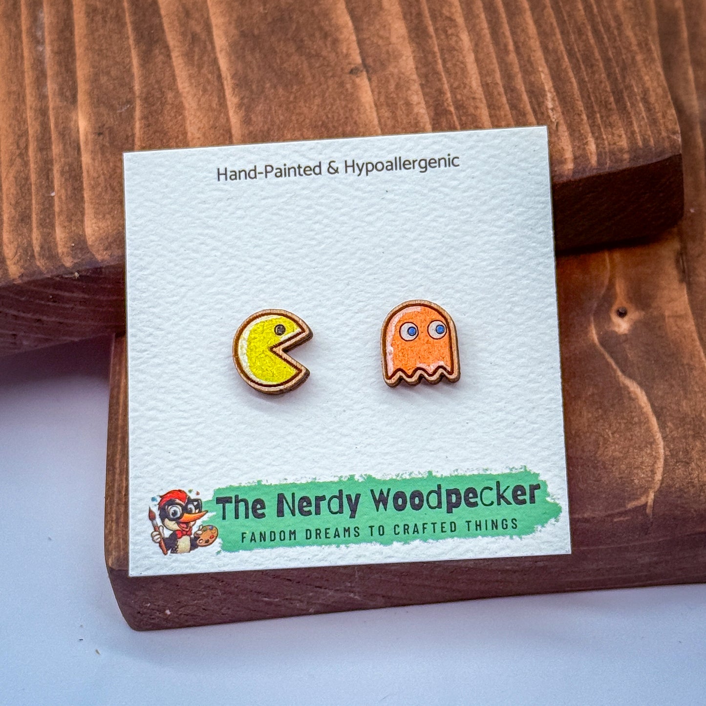 two earrings, one shaped like Pac-Man and the other like a ghost, displayed on a wooden surface.
