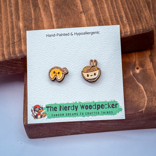 two earrings, one shaped like a fortune cookie and the other a bowl of noodles, presented on a wooden surface.