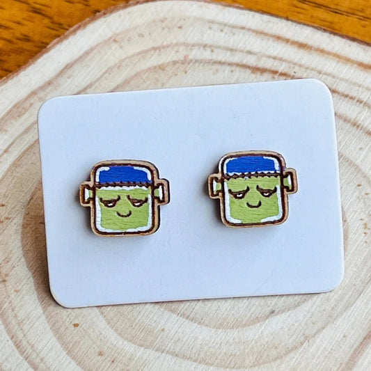 two wooden earrings with cartoon-like faces, each with a blue hat and a green body, placed on a white card.