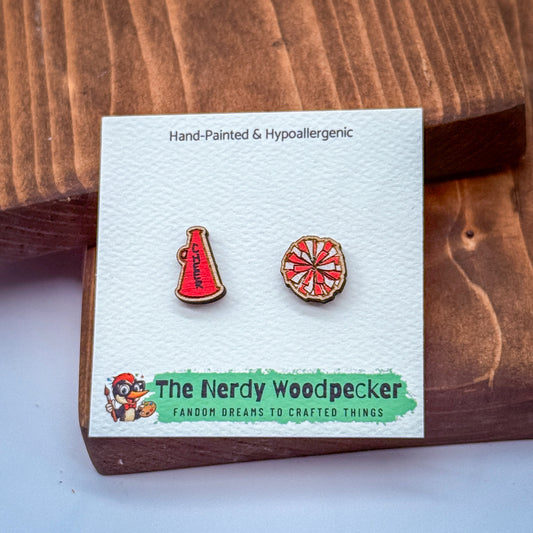 two earrings, one shaped like a megaphone and the other like a pizza wheel, presented in a wooden box with a white card.