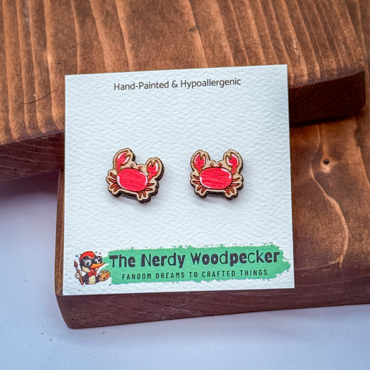 A pair of hand-painted and hypoallergenic earrings in the shape of red crabs, presented on a white card with text and a cartoon character.