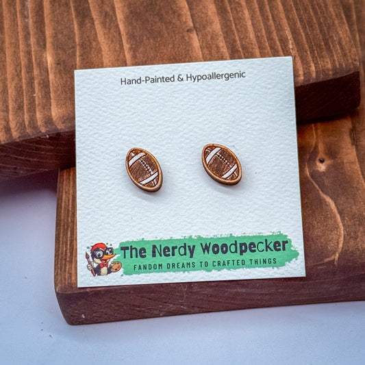 a pair of hand-painted football earrings displayed on a wooden surface, with a white card underneath that has text and a logo.