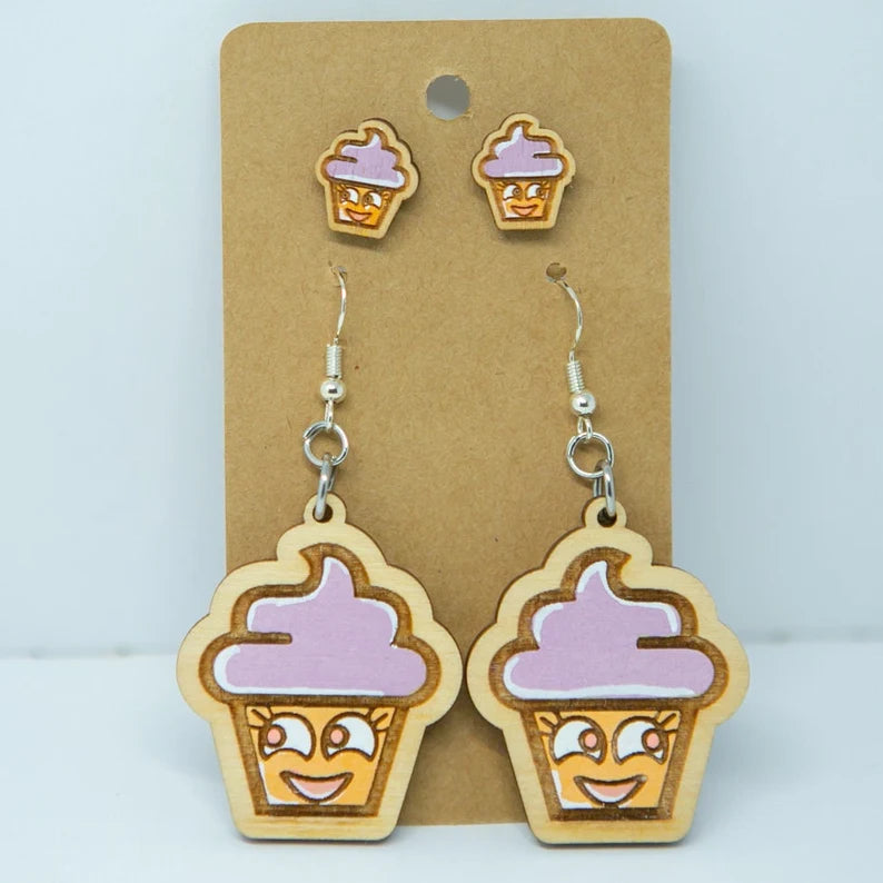 A pair of wooden earrings with cartoon cupcake faces and purple frosting, displayed on a brown card.