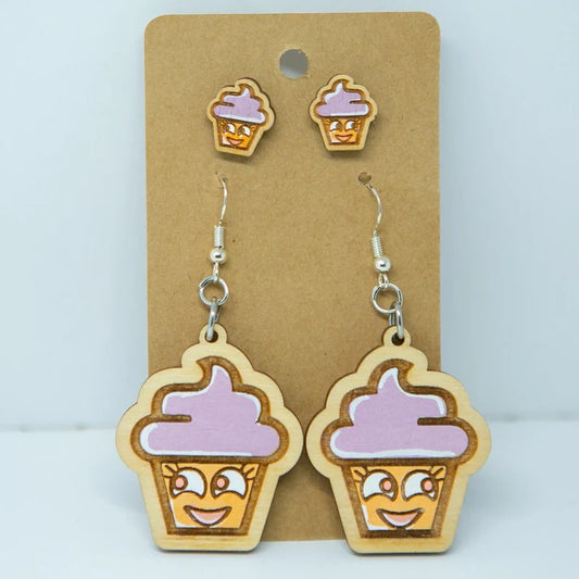 A pair of wooden earrings with cartoon cupcake faces and purple frosting, displayed on a brown card.