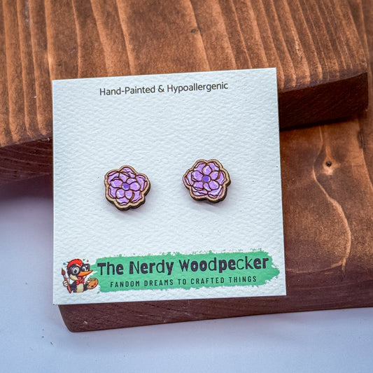 a pair of hand-painted earrings with purple flowers, displayed on a wooden surface. The earrings are part of a product from "The Nerdy Woodpecker", a fan of fantasy and crafted items.