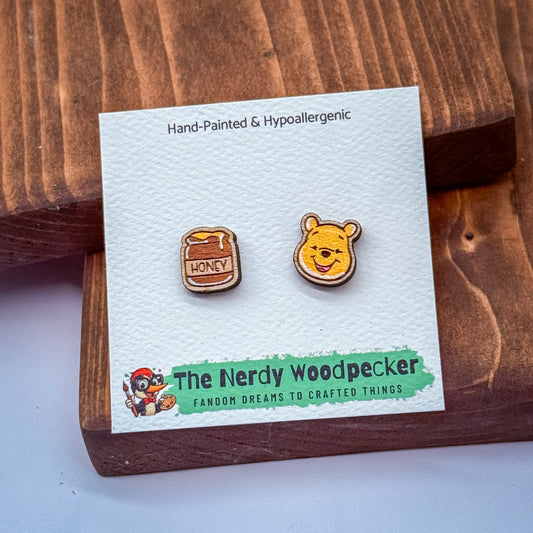 two earrings, one shaped like a honey jar and the other a cartoon character, presented on a wooden surface.