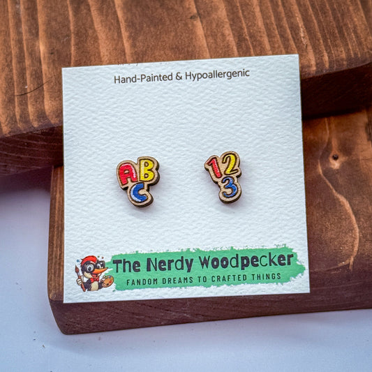 a pair of hand-painted and hypoallergenic earrings with the text "The Nerdy Woodpecker Fandom Dreams to Crafted Things" on a wooden surface.