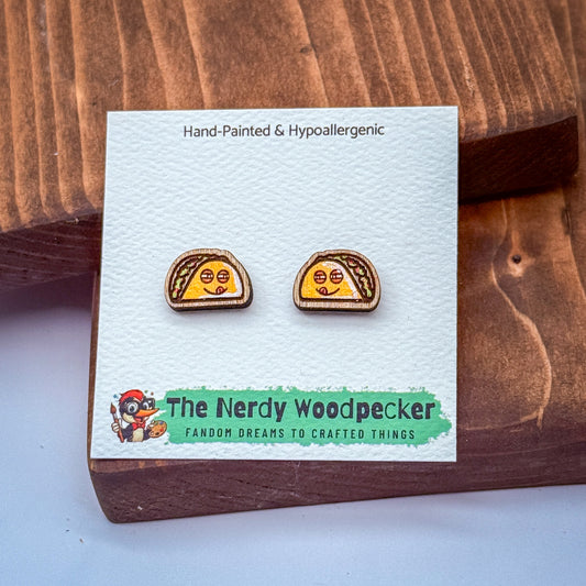 a pair of earrings with a hand-painted design, placed on a wooden surface. The earrings are described as hypoallergenic and are part of a collection by The Nerdy Woodpecker, which features fan-made dreams to crafted things.
