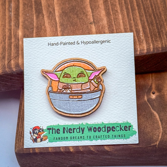 A hand-painted wooden pin depicting a young alien character, likely representing the character Baby Yoda from the Star Wars franchise, is displayed on a white card.